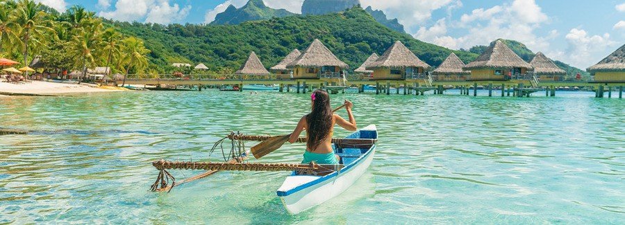 Woman paddling outrigger canoe in Tahiti lagoon with overwater bungalows and mountains