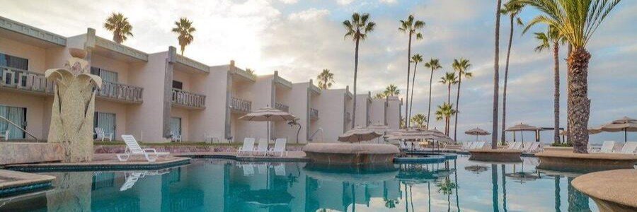 Ensenada beaches at Estero Beach Hotel resort with a calm pool surrounded by palm trees, lounge chairs, and coastal-style buildings, overlooking the Pacific Ocean in Baja California Mexico with a relaxed beachfront setting