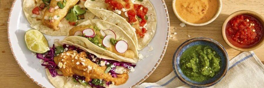 Ensenada beaches local cuisine featuring traditional fish tacos served with fresh toppings, including cabbage, radishes, salsa, and lime, alongside small bowls of sauces, showcasing a classic Baja California seafood dish