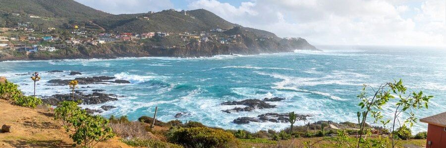 Ensenada beaches at Punta Banda with rugged rocky coastline, waves crashing against dark rocks, and hillside homes overlooking the Pacific Ocean, showing a dramatic coastal landscape in Baja California Mexico