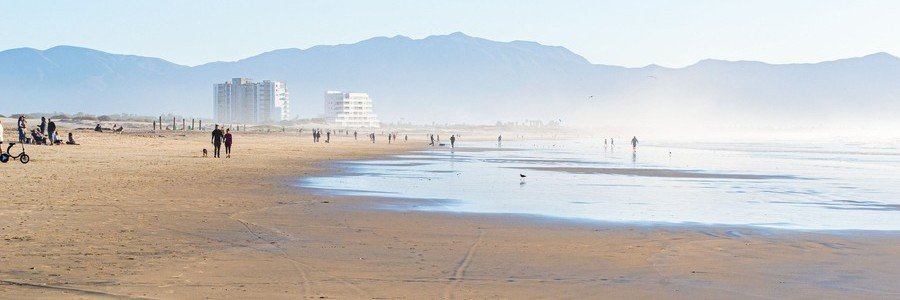Ensenada beaches with a wide sandy shoreline, shallow reflective water, and people walking along the coast, with mountains and buildings in the distance under a soft coastal haze in Baja California Mexico