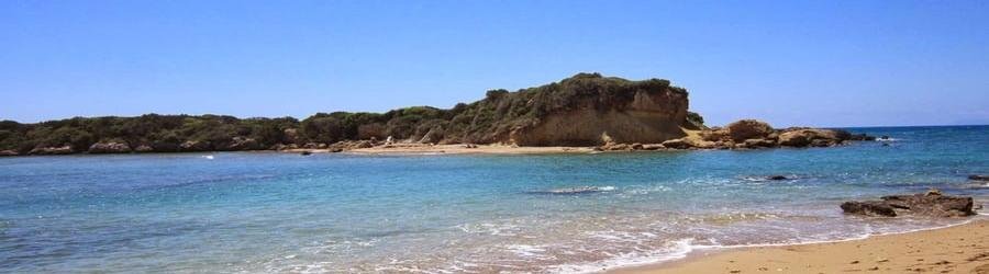 Katakolon beach in Greece with golden sand, rocky outcrop, and clear turquoise sea under a bright blue sky.