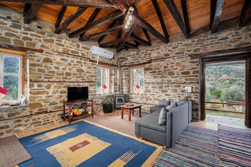 Rustic living room in a stone cottage with exposed wooden beams, gray sofa, and a TV on a wooden stand near a stone wall windowed with a view outside.