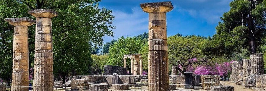 Ancient stone columns and ruins in a sunny park with green trees and purple flowering bushes in the background.