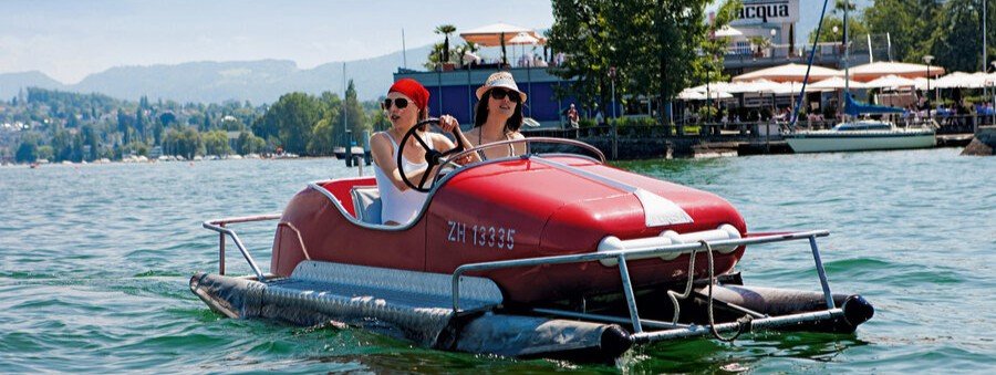 People riding a red pedal boat on Lake Zurich with waterfront cafés and mountains in the background