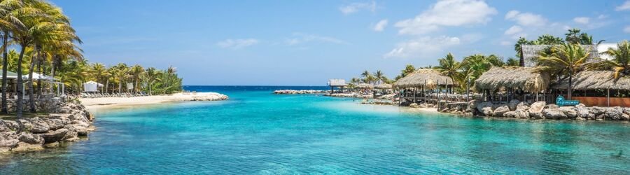 Palm trees and thatched beach bars lining a calm turquoise bay at a seaside lagoon in Curacao.