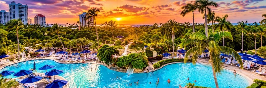 Naples Grande Beach Resort in Naples, Florida, featuring a tropical lagoon pool, waterfalls, palm trees and lush resort grounds at sunset.