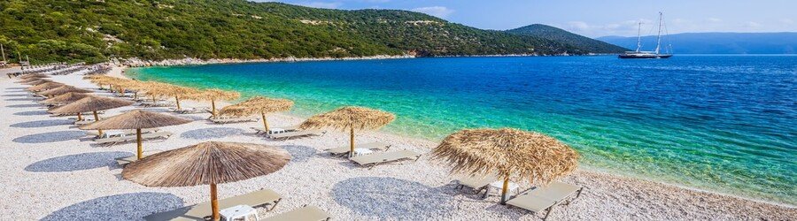 Sunbeds and straw umbrellas lined up along the white-pebble shore of Antisamos Beach in Kefalonia, with bright turquoise water and green hills.