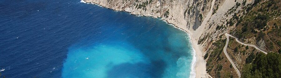 A dramatic aerial view of Myrtos Beach in Kefalonia, showcasing deep blue water, steep cliffs, and a long white-pebble shoreline.