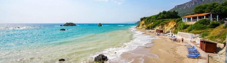 A long sandy beach in Corfu with blue sunbeds, turquoise water, and a small seaside taverna.