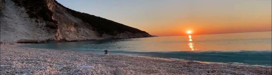 Sunset over Myrtos Beach in Kefalonia with glowing orange light reflecting on the calm sea and white pebbles.