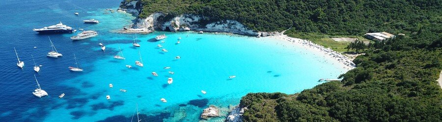 Aerial view of Voutoumi Beach in Antipaxos with bright turquoise water, anchored boats, and lush green hills.