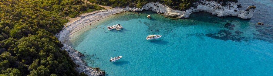 Aerial view of a secluded Antipaxos cove with bright blue water, white rocky shores, and small boats anchored close to the beach.