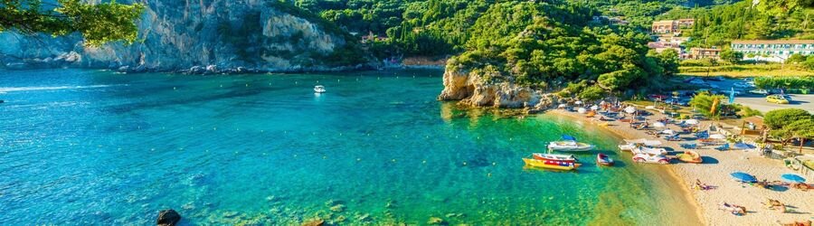 A turquoise bay in Paleokastritsa, Corfu, with lush green hills, boats, and a sandy beach lined with sunbeds.