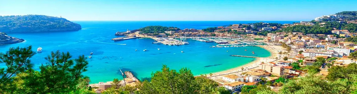 Coastal harbor town in the Balearic Islands with turquoise water, marina, sandy beach, and Mediterranean hillside views