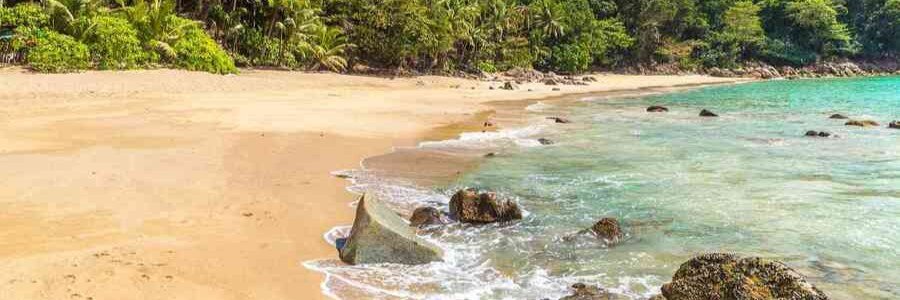 10 Most Beautiful Beaches | Banana Beach, Thailand Banana Beach in Thailand with golden sand, clear turquoise water, shoreline rocks, and dense tropical greenery.