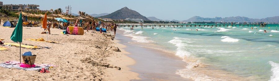 10 Most Beautiful Beaches | Playa de Muro Playa de Muro in Mallorca with soft sand, shallow turquoise water, beach umbrellas, swimmers, and distant mountains.