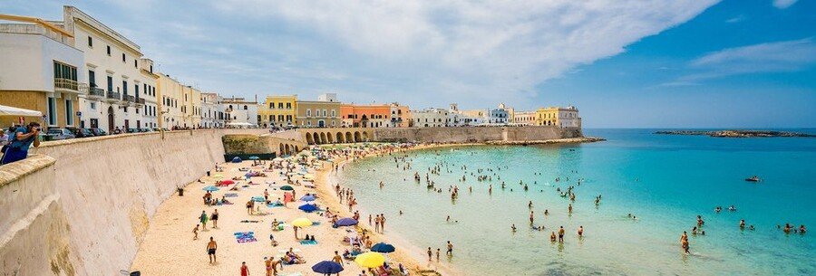 Historic seaside town in Puglia, Italy with colorful buildings, a stone seawall, and people swimming in clear turquoise water