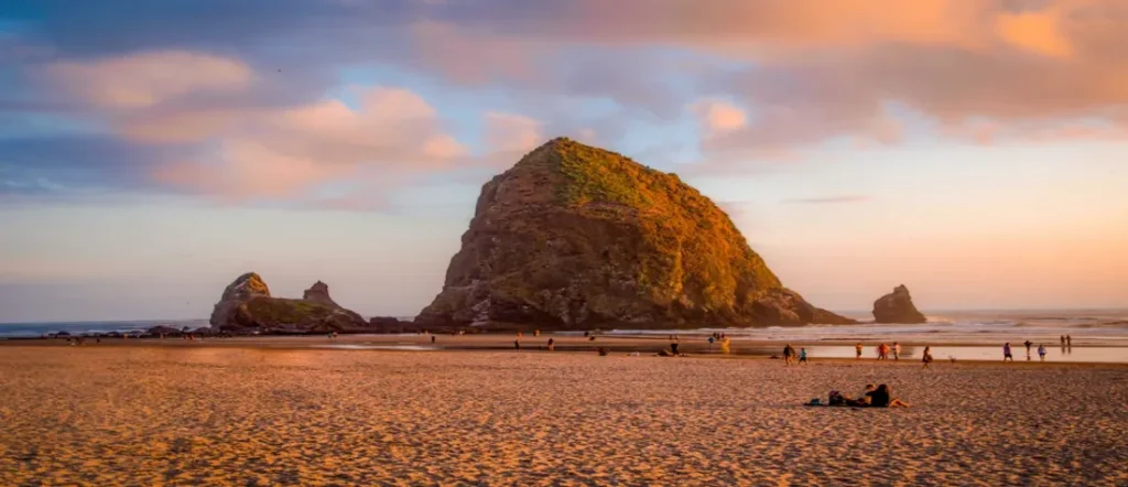 Haystack Rock at sunset on Cannon Beach Oregon with soft golden light and people walking along the shoreline.