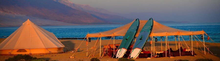 Coastal glamping tents set up along the Socotra shoreline at sunset with kayaks and mountains in the distance.