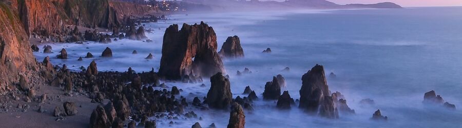 Rocky shoreline at sunset along the Del Norte County coast with sea stacks and misty waves.