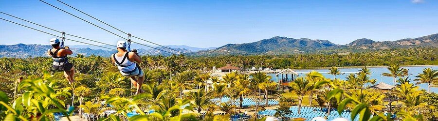 Two people ziplining over the palm-filled landscape and lagoon area at Amber Cove in the Dominican Republic with mountains in the background.