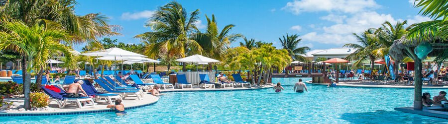 Resort-style pool at the Grand Turk Cruise Center with palm trees, sun loungers, and visitors relaxing under clear blue skies.