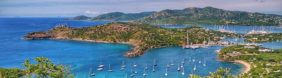 Panoramic view of English Harbour and surrounding green hills in Antigua, featuring turquoise water dotted with sailboats.