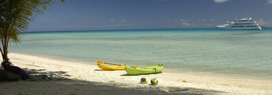 Yellow and green kayaks resting on a white sand beach with calm lagoon water and a boat in the distance in Rangiroa