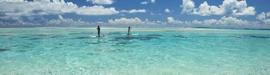 Two people paddleboarding in shallow clear turquoise lagoon water with clouds overhead