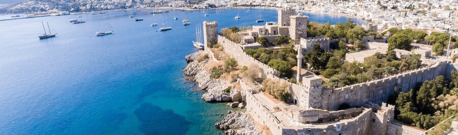 Aerial view of the stone walls and towers of Bodrum Castle on a rocky peninsula surrounded by the bright blue Aegean Sea with sailboats in the harbor.