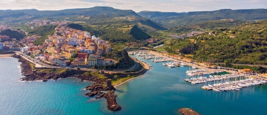 Aerial panorama of Castelsardo, Sardinia, featuring its colorful hillside buildings, medieval castle, and a busy marina filled with white sailboats on bright blue water. Near Cagliari Port Sardinia Italy