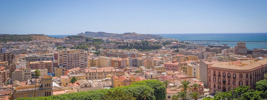 Panoramic cityscape of Cagliari, Sardinia, showing densely packed colorful buildings, historic architecture, and the Mediterranean coastline under a clear blue sky. Start your Cagliari Port Day here.