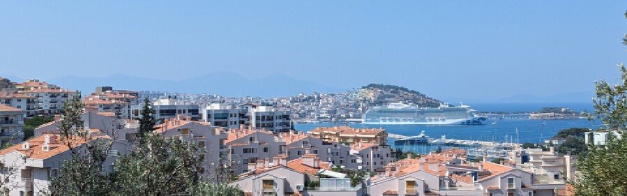 Scenic view of Kuşadası city rooftops with a large cruise ship docked in the Aegean Sea harbor.