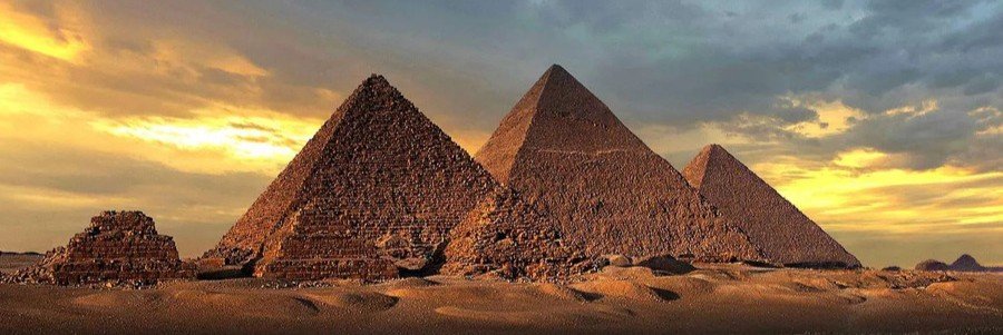 The three Great Pyramids of Giza at sunset, with golden light hitting the stone structures and a dramatic sky in the background.