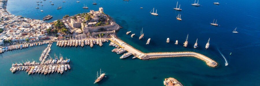 An aerial drone view of Bodrum Port showing the historic Castle of St. Peter, the long pier with docked boats, and the white-washed buildings of the city lining the turquoise Aegean coast.