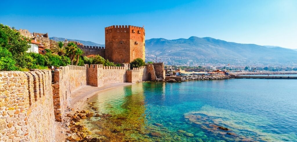 A wide-angle view of the historic Antalya port area in Alanya, featuring the iconic octagonal Red Tower (Kızıl Kule) and ancient stone fortress walls meeting the clear, turquoise Mediterranean sea under a bright blue sky.