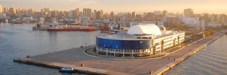 An aerial golden-hour view of the modern, white-domed Alexandria Port terminal building situated on a long pier, with cargo ships and the city’s dense urban skyline in the background.