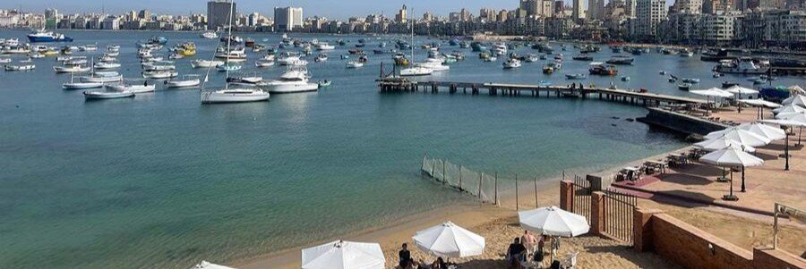 A panoramic view of the Eastern Harbor on the Alexandria Waterfront, filled with white sailboats and fishing boats, with the city skyline and a beach club in the foreground.
