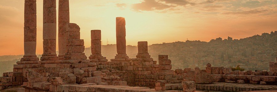 The ancient stone columns of the Temple of Hercules at the Amman Citadel in Jordan, silhouetted against a warm orange sunset with the city in the background.