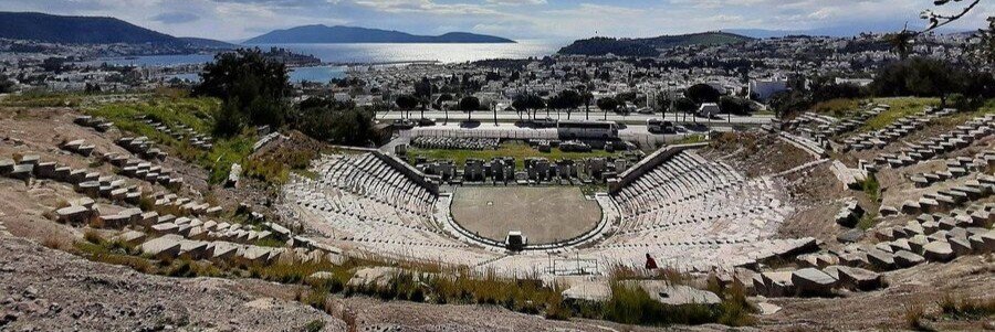 Wide panoramic view looking down from the stone seating of the Ancient Theatre of Halicarnassus toward the stage, with the city of Bodrum and the Aegean Sea in the background.
