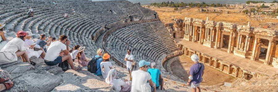 ancient-theatre-hierapolis-pamukkale-turkey | Beach Travel Destinations A wide panoramic view from the upper tiers of the ancient Roman theater at Hierapolis, showing tourists sitting on stone seats overlooking a massive semi-circular cavea and a highly detailed, two-story stone stage building with ornate columns and niches under a clear blue sky.