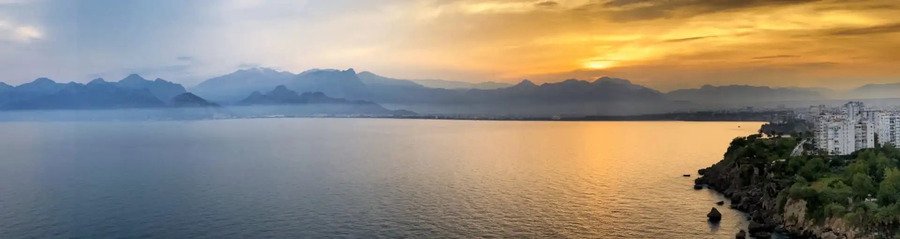antalya-bay-sunset-panorama-cliffs | Beach Travel Destinations A panoramic sunset view over the Antalya Bay in Turkey, featuring the shimmering Mediterranean Sea, a dramatic silhouette of the Taurus Mountains in the distance, and modern white apartment buildings perched on the rugged coastal cliffs to the right.