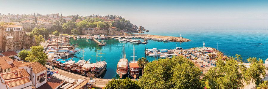 antalya-kaleici-old-town-marina-panorama | Beach Travel Destinations A high-angle panoramic view of the historic Kaleiçi Old Town Marina in Antalya, Turkey, featuring numerous tour boats and yachts anchored in a turquoise crescent-shaped bay, surrounded by traditional Ottoman-style houses with red-tiled roofs and lush green trees under a clear sky.