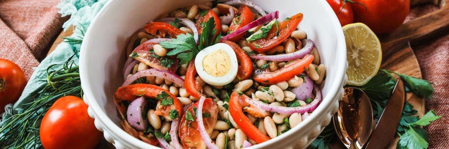 antalya-piyaz-bean-salad-tahini | Beach Travel Destinations A top-down close-up view of a white ceramic bowl filled with Antalya Piyazı, featuring white beans, sliced red onions, fresh parsley, and wedges of ripe tomato, topped with a perfectly halved hard-boiled egg and a creamy tahini dressing.