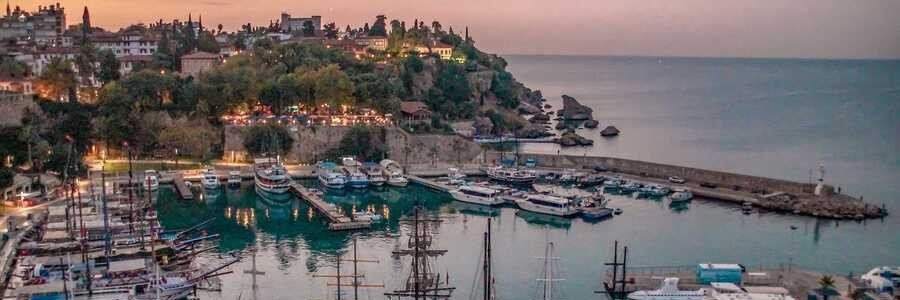 A panoramic evening view of the historic Antalya port in the Kaleiçi district, showing numerous yachts and excursion boats docked in the calm marina with the glowing lights of the old city and surrounding cliffs in the background.
