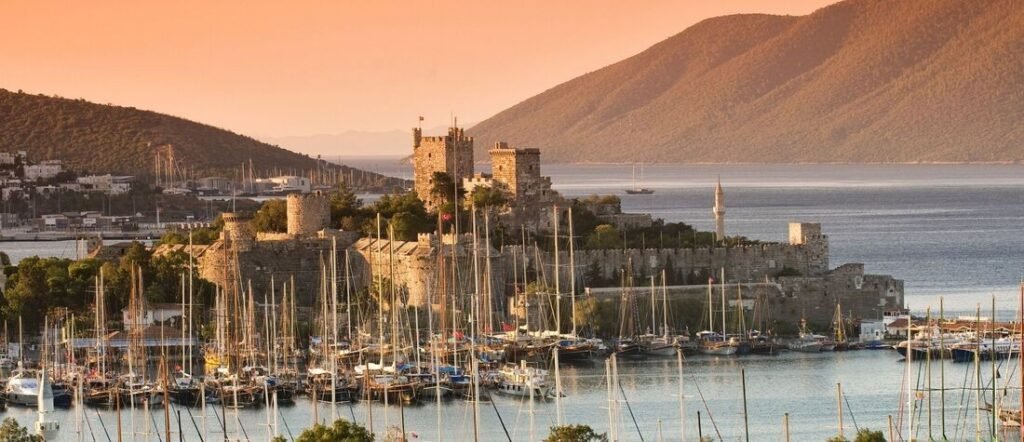 A wide panoramic view of Bodrum Port at sunset, featuring the historic medieval Castle of St. Peter, numerous sailboats and yachts docked in the harbor, and rolling green hills in the background under a soft orange sky.