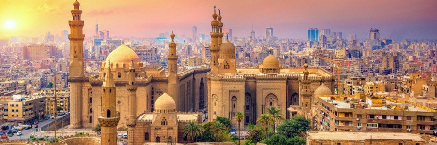 A high-angle panoramic view overlooking the historic Sultan Hassan Mosque and Al-Rifa'i Mosque complexes under a warm golden sunset in Cairo, Egypt.