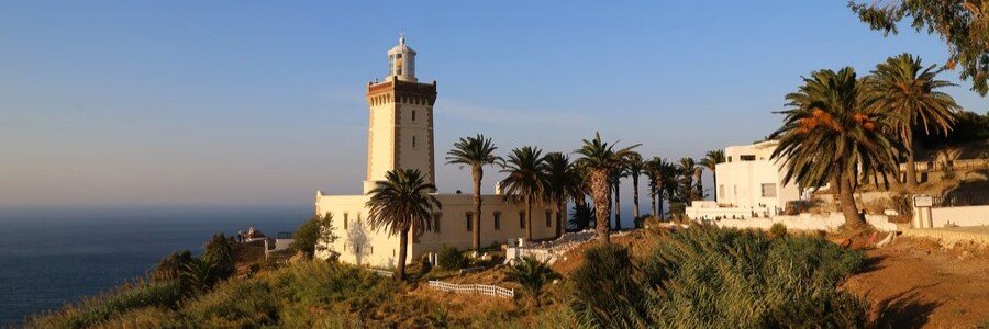 The historic yellow-and-white Cape Spartel Lighthouse in Tangier, Morocco, surrounded by palm trees and coastal greenery.