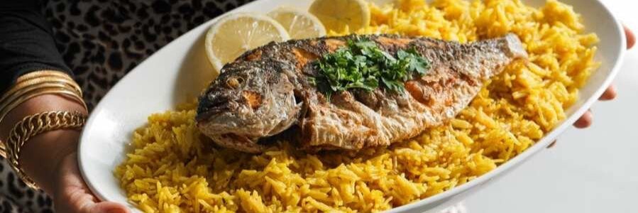 A close-up view of an oval platter filled with golden yellow sayadieh rice topped with a whole fried fish garnished with fresh parsley and lemon slices.